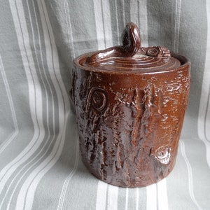 May include: A brown ceramic jar with a tree bark texture and a lid shaped like a tree branch. The jar is sitting on a striped fabric.