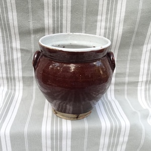 May include: A brown ceramic pot with two handles and a white glazed interior. The pot is sitting on a striped fabric background.
