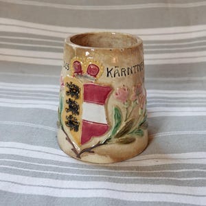 Puede incluir: Una taza de cerámica con fondo marrón y un diseño pintado de un escudo de armas con una corona, un escudo rojo y blanco, y el texto "KÄRNTNER" en negro. La taza está decorada con hojas verdes y flores rosas.