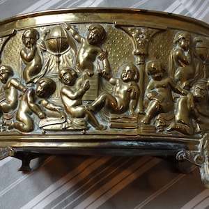 Può includere: Un vaso da fiori ovale in ottone con tre piedi. Il vaso è decorato con un rilievo di cherubini che suonano musica e tengono un globo.