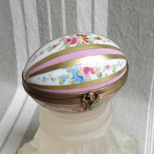 Könnte beinhalten: Eine porzellanene, eiförmige Schmuckschatulle mit einem Scharnierdeckel. Die Schachtel ist mit rosa und goldenen Streifen und handgemalten Blumendesigns in den Farben Rosa, Blau und Grün verziert.