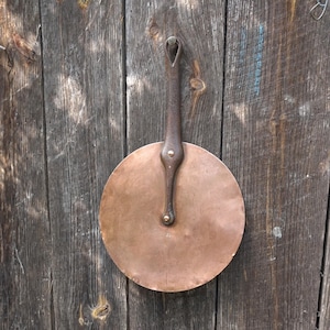 Peut inclure: Une poêle en cuivre avec une longue poignée en fer noir. La poêle est accrochée à un mur en bois.