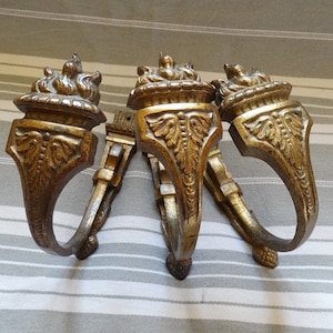 May include: Three ornate gold-toned metal curtain tiebacks with a decorative design featuring a flame-like motif at the top. The tiebacks are curved and have a decorative design throughout.