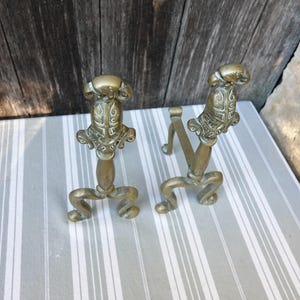 May include: A pair of antique brass fireplace andirons with ornate designs. The andirons are shaped like stylised figures with a decorative top.