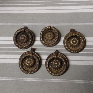 May include: Five antique brass drawer pulls with a circular design. Each pull features a central floral motif surrounded by an ornate, textured ring and a hinged handle. The pulls are mounted on small leather tabs.