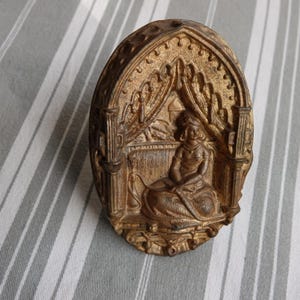 Puede incluir: Placa ovalada de metal dorado antiguo que presenta una figura sentada dentro de un marco arqueado. La figura está representada en un estilo clásico detallado, con elementos arquitectónicos ornamentados y cortinajes.