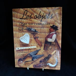 Puede incluir: Un libro titulado "Les objets de nos campagnes" con varias herramientas antiguas y una figura de madera. La portada presenta un fondo de textura tejida. Las herramientas incluyen tijeras, una flauta y un plato en forma de corazón.