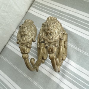 May include: A pair of ornate gold-tone metal hooks with lion head designs. The hooks are attached to a wall and have a decorative scrollwork design.