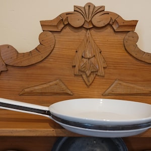 May include: A white frying pan with a black and white handle. The pan has a dark gray band around the rim. The pan is on a wooden surface with a carved wooden headboard in the background.
