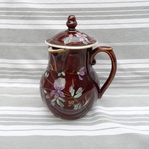 以下が含まれることがあります： 蓋付きの茶色の陶器のピッチャーで、紫、緑、金の色の花柄で飾られています。ピッチャーには取っ手と注ぎ口があります。