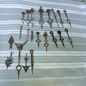 Peut inclure: Une collection d'aiguilles d'horloge anciennes de formes et tailles variées, en métal vieilli. Les aiguilles présentent des motifs ornés, notamment des cercles, des croix et des pointes. Elles sont disposées sur un tissu rayé gris clair et blanc.