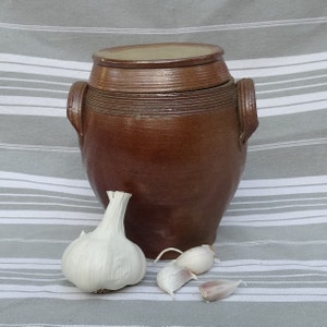 May include: A brown ceramic jar with a lid, sitting on a striped fabric surface. There are four white garlic bulbs next to the jar.