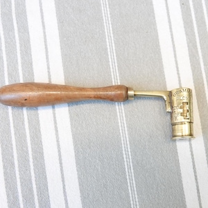 May include: A brass tool with a wooden handle, used for measuring lead shot. The tool has a dial with markings in grams and a small opening for dispensing the shot.