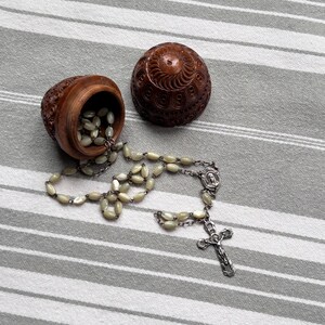 Könnte beinhalten: Ein brauner, geschnitzter Holzbehälter mit passendem Deckel, der geöffnet dargestellt ist, wobei ein Rosenkranz herausfällt. Der Rosenkranz besteht aus hellen, ovalen Perlen, silberfarbenen Metallgliedern, einer kleinen Medaille und einem Kruzifix.