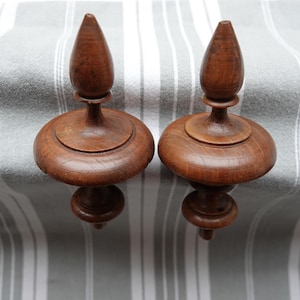 Op de afbeelding: Twee houten ornamenten met een donkerbruine afwerking. Elk ornament heeft een afgeronde basis, een centrale steel en een conische top. De ornamenten worden gepresenteerd op een grijs en wit gestreept oppervlak.