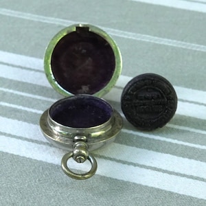 May include: An open, silver-toned, round container with a purple velvet interior, next to a dark, round object with embossed text. The container has a hinged lid and a small ring attached. The objects are on a striped background.