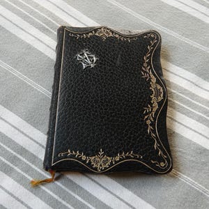May include: A black leather-bound book with a textured surface, featuring gold-colored floral filigree detailing along the right edge and a silver-toned metallic emblem with an intricate monogram design attached to the upper left corner.