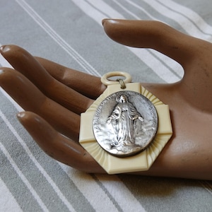 May include: A vintage religious pendant featuring a silver medallion of a saint figure, set on a cream-colored, geometric base. The pendant has a ring for hanging. The medallion is detailed with text around the edge.