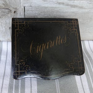 Könnte beinhalten: Schwarze Zigarettenetui mit goldfarbenem Schriftzug und dekorativen Eckakzenten. Das Wort "Cigarettes" ist in eleganter Schrift geschrieben. Die Hülle ist rechteckig mit einer gebogenen Unterkante.