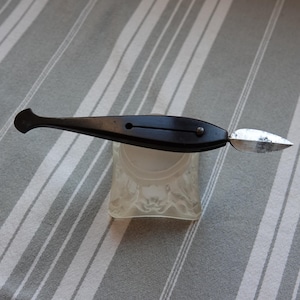 May include: A vintage black pen with a silver nib, resting on a clear glass inkwell. The pen has a sleek, elongated design with a rounded handle. The inkwell is square with a decorative pattern. The background is a gray and white striped fabric.