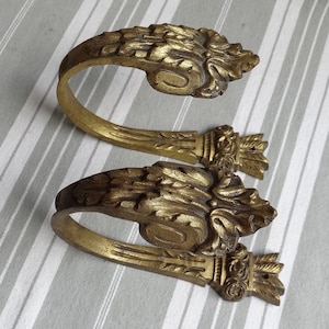 May include: Two gold-toned metal curtain tiebacks with ornate floral designs. The tiebacks are shaped like a hook and have a decorative base.