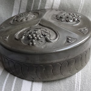 Puede incluir: Una caja redonda de peltre con un intrincado diseño de uvas y hojas. La caja tiene una tapa con bisagras y está decorada con un diseño en relieve. La caja está firmada "Made in England".
