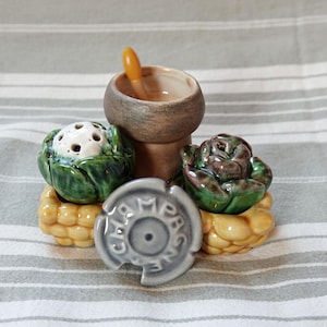 Può includere: Un set di saliere e pepiere in ceramica a forma di verdure. Le saliere sono verdi e bianche e si trovano su una base gialla. Il set include anche un piccolo contenitore in ceramica marrone con un cucchiaio di legno.