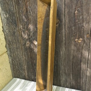 Könnte beinhalten: Ein langer, schmaler Holztrough mit leicht gekrümmter Form. Der Trog ist aus Naturholz in hellbrauner Farbe und hat ein abgenutztes, rustikales Aussehen.