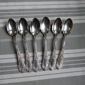 May include: Six silver spoons with ornate, detailed handles are arranged on a gray and white striped surface. The spoons have a reflective, polished finish, and the handles feature a scrolling floral design, suggesting a vintage or antique style.