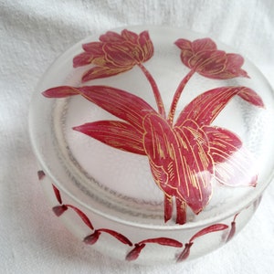 May include: A clear glass jar with a red and gold floral design. The jar has a lid and is decorated with a repeating pattern of red flowers around the rim.