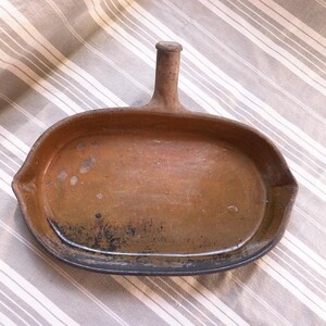 Puede incluir: Una sartén rústica de terracota de forma ovalada con un asa. La sartén tiene un interior marrón oscuro y un borde negro. El asa es cilíndrica y se eleva desde el centro de la sartén. La sartén probablemente se utiliza para cocinar o servir.