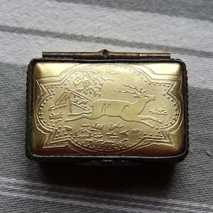 May include: A rectangular, hinged brass box with a gold-colored lid. The lid features an embossed design of a deer leaping across a landscape with trees and a fence. The box has a dark, possibly tarnished, border.