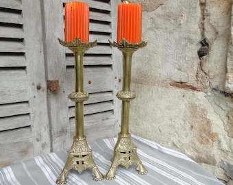 May include: Two antique brass candle holders with orange candles. The candle holders have ornate bases and detailed designs. The candles are cylindrical and have a ribbed texture. The background includes a stone wall and a wooden shutter.