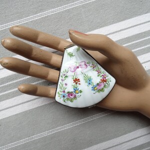May include: A white porcelain trinket box shaped like a seashell with a floral design in shades of pink, blue, green, red, and yellow. The box has a brass hinge and a floral design on the lid.