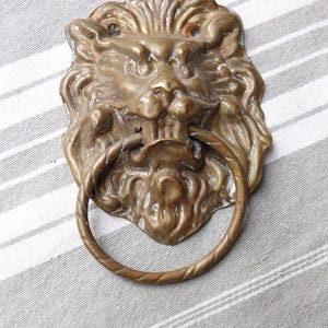 Op de afbeelding: Een messing deurklopper in de vorm van een leeuwenkop met een ringvormige handgreep. De deurklopper is gedetailleerd met een getextureerde manen en een woeste uitdrukking.