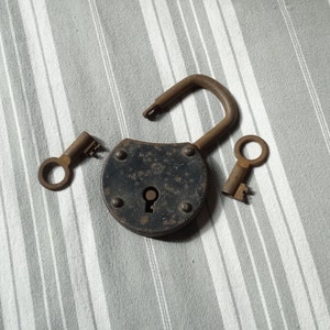 May include: A vintage padlock with a black metal body and a keyhole. Two keys are included, one with a round handle and one with a more traditional keyhole.