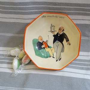 Puede incluir: Un plato de cerámica octogonal blanco con un borde naranja. El plato presenta una ilustración de dibujos animados de dos hombres, uno sirviendo al otro una botella de licor en una bandeja. El texto "His master's vice" está escrito en el plato.