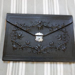 Könnte beinhalten: Eine rechteckige Dokumentenmappe aus schwarzem Leder mit einem geprägten Ranken- und Blattmuster auf der Vorderklappe. In der Mitte der Klappe befindet sich ein silberfarbener Metallverschluss mit einem kleinen Schlüsselloch.