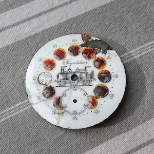 May include: Antique white clock face with a black and white train illustration and the word "Regulateur". The face has red and yellow accents and is cracked in several places. Numbers are visible around the edge.