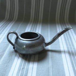Può includere: Una piccola lampada a olio antica in peltro con un corpo arrotondato, un manico e un lungo beccuccio curvo. La lampada ha un aspetto scuro e invecchiato, che suggerisce che sia vintage. Lo sfondo è un tessuto a righe.