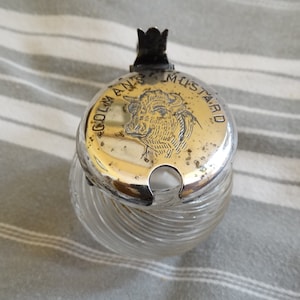 May include: A vintage glass mustard jar with a silver lid. The lid features a bull's head design and the text "COLMAN'S MUSTARD" around the edge.