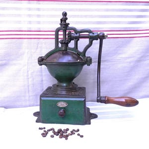May include: Antique green and black coffee grinder with a wooden handle. The grinder has a round top and a square base. Coffee beans are scattered in front of the grinder.