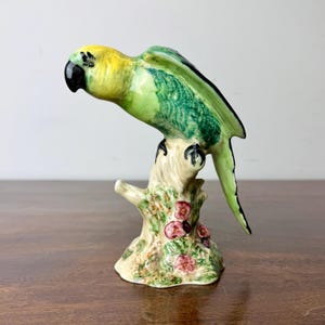 May include: A ceramic parrot ornament, predominantly green and yellow, perched on a tree branch. The parrot has a black beak and feet. The branch is beige with pink flowers. A decorative item, suitable for display.