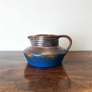 May include: A brown and blue ceramic jug with a wide base and a rounded body. The jug has a brown glaze with a blue glaze on the bottom.
