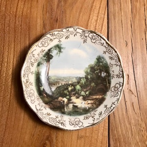 May include: A white ceramic plate with a gold rim and a floral design. The centre of the plate features a painted landscape scene with a bridge, trees, and a river.