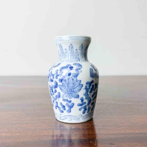May include: A small white ceramic vase featuring a hand-painted blue floral pattern. The design includes a central lotus-like flower surrounded by scrolling foliage, with a repeating leaf motif around the neck and wavy lines at the base.