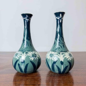 May include: Two matching ceramic vases with a narrow neck and a bulbous base. The vases feature a dark teal and sage green glaze with hand-painted white daisy-like flowers and dark green foliage patterns. Both pieces have a visible crackle finish.