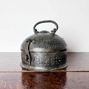 May include: An antique, ornately engraved, dome-shaped metal box with a hinged lid and a carrying handle. The box is made of dark metal with intricate patterns and a silver-coloured finish.
