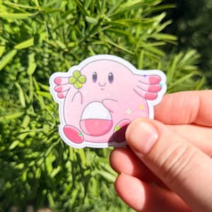 K&ouml;nnte beinhalten: Ein rosa Aufkleber mit einer Zeichentrickfigur mit wei&szlig;em Bauch, die ein vierbl&auml;ttriges Kleeblatt h&auml;lt. Der Aufkleber hat ein glitzerndes Finish und ist vor einem Hintergrund aus gr&uuml;nen Bl&auml;ttern angebracht.