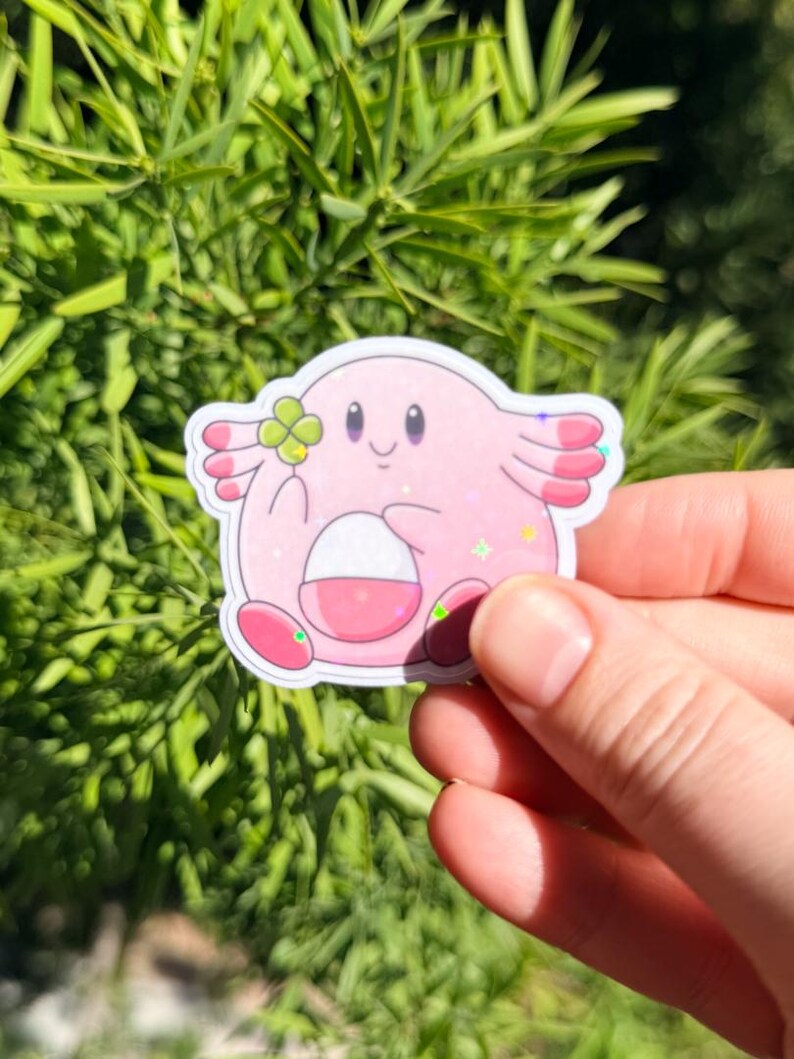 K&ouml;nnte beinhalten: Ein rosa Aufkleber mit einer Zeichentrickfigur mit wei&szlig;em Bauch, die ein vierbl&auml;ttriges Kleeblatt h&auml;lt. Der Aufkleber hat ein glitzerndes Finish und ist vor einem Hintergrund aus gr&uuml;nen Bl&auml;ttern angebracht.